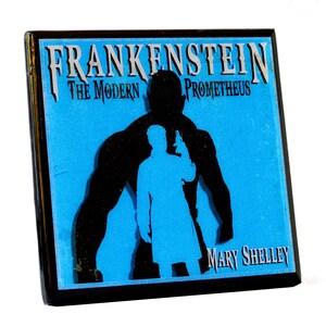 May include: Square pin featuring the title "FRANKENSTEIN The Modern Prometheus" with a silhouette of a figure holding a gun, and a muscular figure in black on a blue background. The author's name, Mary Shelley, is also displayed.