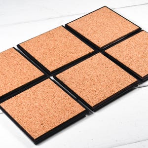 May include: Six square cork coasters with black frames. The coasters are arranged in a staggered pattern.