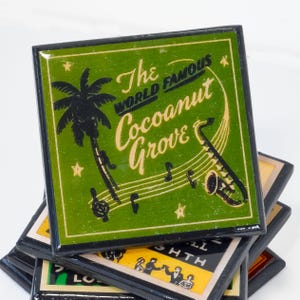 May include: A stack of four green and black coasters with a black border. The top coaster features a palm tree, musical notes, a saxophone, and the text "The World Famous Coconut Grove".