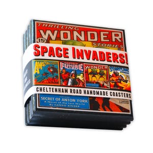 May include: Set of six square coasters featuring vintage comic book covers with a space theme. The coasters are made of cork and have a black border. The text on the coasters reads "Space Invaders!" and "Cheltenham Road Handmade Coasters".