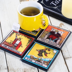 May include: A yellow mug with tea rests on four square tarot card coasters. The coasters feature colorful illustrations and the titles "The Magician," "The Fool," "The Emperor," and "Pentacles." A tea bag is visible.