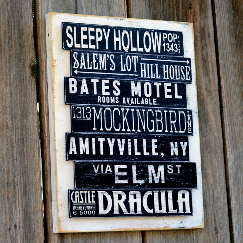 Pu&ograve; includere: Un cartello di legno bianco con lettere nere che elencano luoghi famosi di film horror. Il cartello recita "Sleepy Hollow Pop. 1343", "Salem's Lot Hill House", "Bates Motel Rooms Available", "1313 Mockingbird Lane", "Amityville, NY", "Via Elm St" e "Castle Transylvania 6-5000 Dracula".