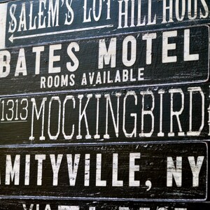 Pu&ograve; includere: Un cartello in bianco e nero con il testo "SLEEPY HOLLOW", "SALEM'S LOT HILL HOUSE", "BATES MOTEL", "ROOMS AVAILABLE", "1313 MOCKINGBIRD", "MITYVILLE, NY" e "VIA ELM ST".