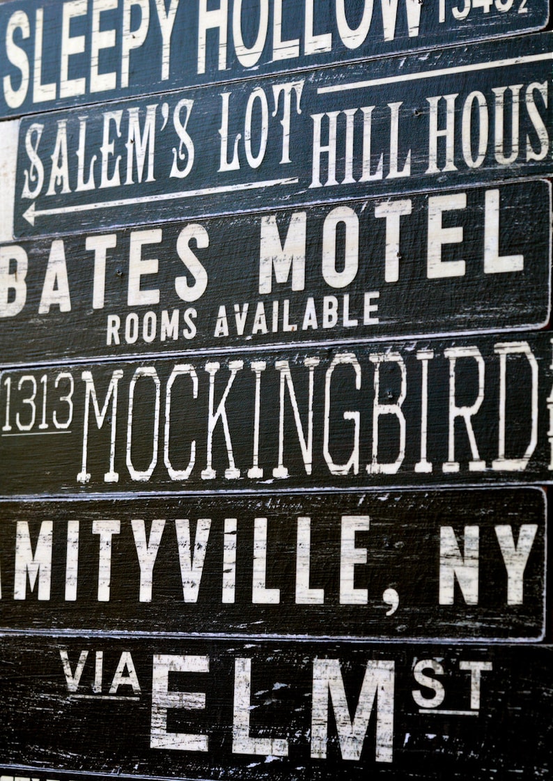 Pu&ograve; includere: Un cartello in bianco e nero con il testo "SLEEPY HOLLOW", "SALEM'S LOT HILL HOUSE", "BATES MOTEL", "ROOMS AVAILABLE", "1313 MOCKINGBIRD", "MITYVILLE, NY" e "VIA ELM ST".