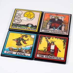 May include: Four square coasters with tarot card designs. The coasters feature images of "Ace of Pentacles", "The Emperor", "The Fool", and "The Magician". Each coaster has a black border and vibrant colors.