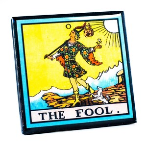 May include: A square magnet featuring a tarot card illustration. The card depicts a figure in colorful clothing with a dog, a sun, and the text "THE FOOL" against a yellow and blue background. The magnet has a black border.