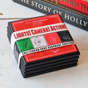 May include: Set of four black coasters with a red, white and green design. The coasters feature a vintage illustration of a film camera and the text "LIGHTS! CAMERA! ACTION!" and "CHELTENHAM ROAD HANDMADE COASTERS".