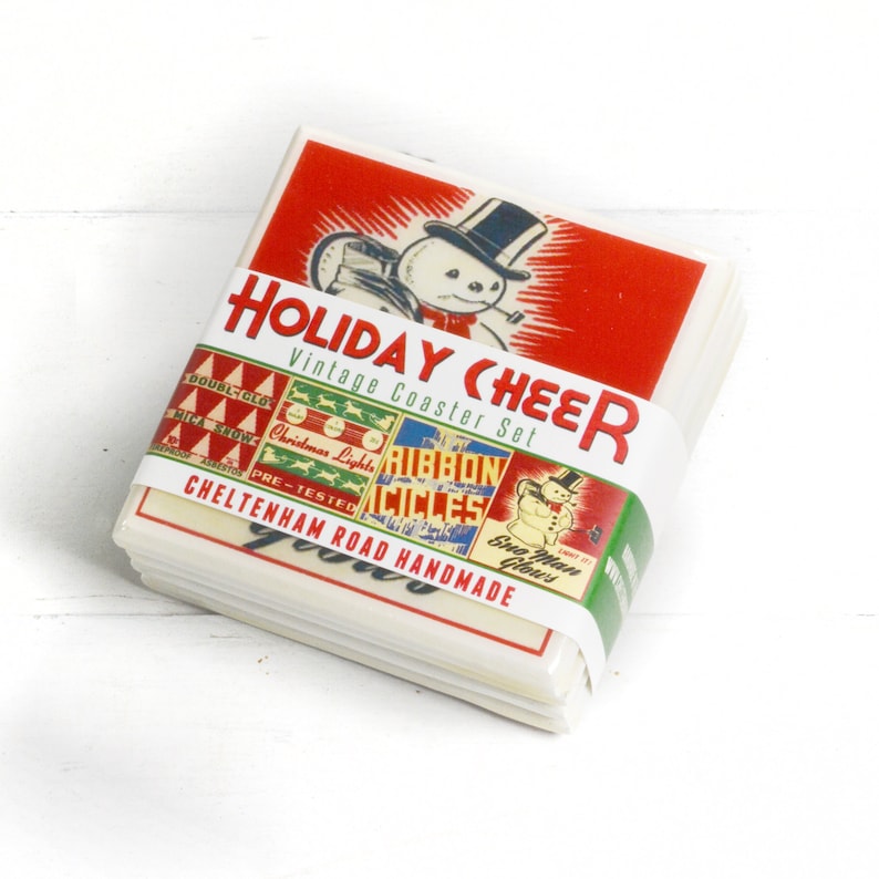 May include: Set of four vintage-style coasters featuring holiday-themed illustrations. The coasters are made of cardboard and feature a snowman, Christmas lights, ribbon, and a snowman with a red scarf. The coasters are packaged together with a red and white label that reads "Holiday Cheer Vintage Coaster Set".