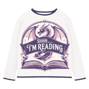 May include: White long-sleeved top featuring a purple and pink dragon design. The design includes a dragon, wings, and an open book, with the text "SHHH... I'M READING" in a banner. The top has navy blue trim.