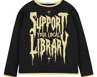 May include: Black long-sleeve shirt with the text "SUPPORT YOUR LOCAL LIBRARY" in a dripping, gothic-style font. The text is in a light tan color, with a matching trim around the collar and cuffs. Two small starbursts are above and below the text.