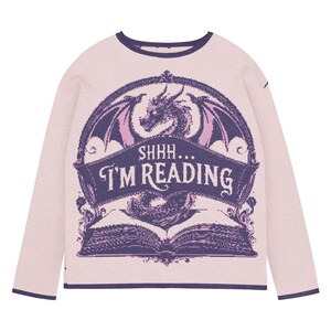 May include: A pale pink long-sleeved sweatshirt with a dark blue trim. It features a purple dragon design and the text "SHHH... I'M READING" above an open book. The design is framed by a decorative border.
