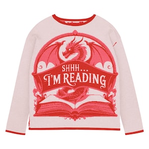May include: A pale pink long-sleeved top with red trim. It features a red dragon design with the text "SHHH... I'M READING". The dragon is set against a pink background with an open book, perfect for book lovers.