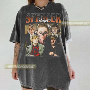 May include: Dark gray t-shirt featuring a graphic of the name "Spencer Wright" in orange and white, with a collage of faces. The shirt has a vintage, worn look. The text "NEWTRENDINGCROCHET" is printed on the shirt.