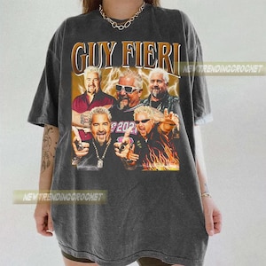 Pode incluir: T-shirt cinza escuro com um gráfico de Guy Fieri e o texto "GUY FIERI" em ouro e castanho. O design inclui várias imagens de Guy Fieri. A camisa tem um estilo vintage.