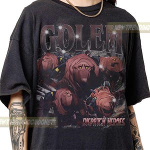 Könnte beinhalten: Dunkelgraues T-Shirt mit einer Grafik von rotbraunen Golems und dem Wort "GOLEM" darüber. Der Text "DISPATCH HEROES" steht darunter. Das T-Shirt hat kurze Ärmel und einen Rundhalsausschnitt.