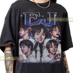 May include: Dark gray t-shirt featuring a collage of images of a person with short dark hair, a microphone, and the letters "EJ" in a blue and white design. The shirt has a vintage, distressed look.