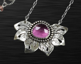 May include: A silver necklace featuring a flower-shaped pendant with a large, faceted, pink gemstone at its center. The pendant has intricate, etched details and is suspended from a delicate chain. The necklace is displayed against a dark background.