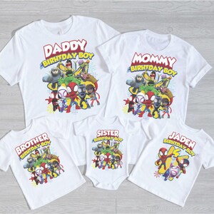 May include: White t-shirts and a onesie featuring a colorful graphic of superheroes. The shirts have text that reads "Daddy Birthday Boy", "Mommy Birthday Boy", "Brother Birthday Boy", "Sister Birthday Boy", and "Jaden Birthday Boy".