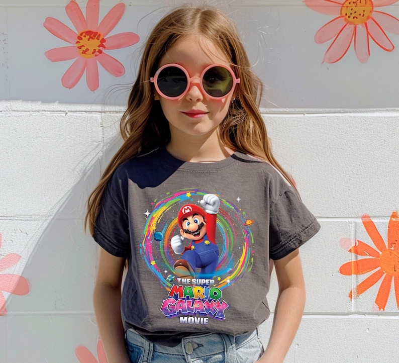 May include: A charcoal gray t-shirt featuring a colorful graphic of Mario from "The Super Mario Galaxy Movie." The design includes Mario with his arm raised, surrounded by a rainbow-colored swirl. The movie title is displayed below the character.