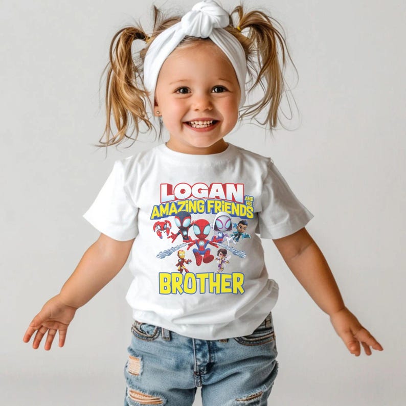 May include: White t-shirt with a colorful graphic featuring the text "LOGAN AND AMAZING FRIENDS BROTHER" and cartoon characters. The shirt is worn by a young child with a white headband and blue jeans.
