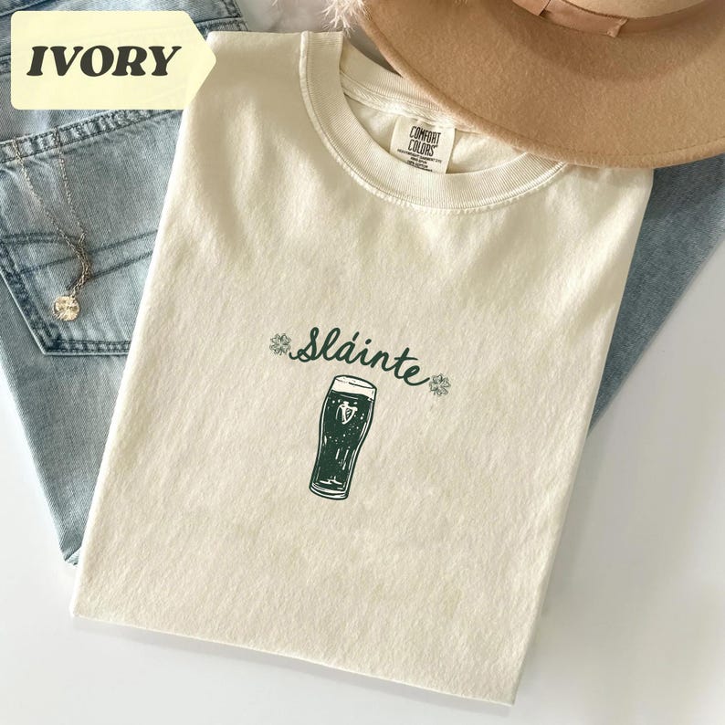K&ouml;nnte beinhalten: Elfenbeinfarbenes T-Shirt mit dem Wort "Sl&aacute;inte" und einer gr&uuml;nen Bierkrug-Grafik. Das Shirt liegt gefaltet auf einer blauen Jeans und einem beigen Hut. Eine silberne Halskette ist sichtbar.