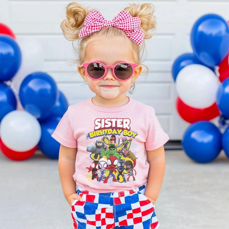 May include: A young child wearing pink sunglasses, a pink t-shirt with the words "Sister Birthday Boy" and a graphic of superheroes. The child is also wearing a pink and white checkered bow and red, white, and blue checkered shorts.
