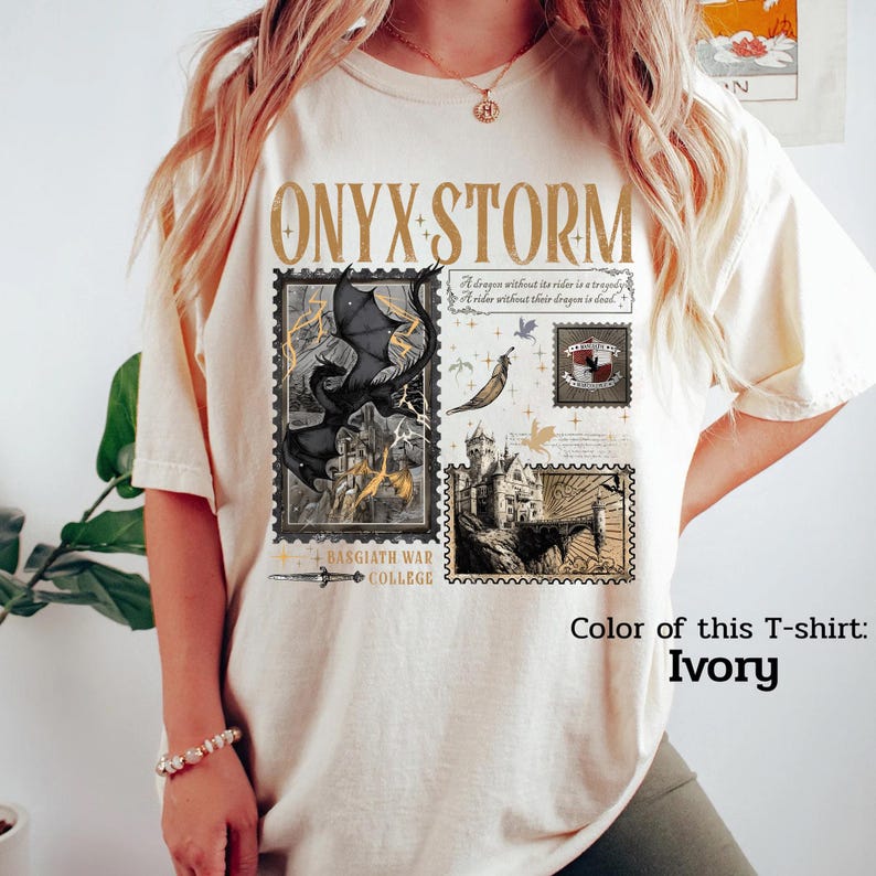 May include: Ivory-colored t-shirt featuring the text "ONYX STORM" in gold lettering. The shirt displays a design with postage stamp-style illustrations of a dragon, castle, and other fantasy elements. The text "BASGIATH WAR COLLEGE" is also present.