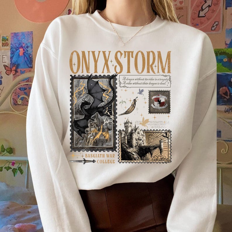 May include: White sweatshirt with the text "ONYX STORM" in gold lettering. The design includes postage stamp-style illustrations of a dragon, castle, and text. The words "BASGIATH WAR COLLEGE" are also printed on the sweatshirt.