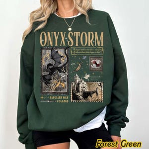 May include: Forest green sweatshirt with the text "ONYX STORM" in gold lettering. The design includes illustrations of a dragon, a castle, and stamps. The text "BASGIATH WAR COLLEGE" is also present.