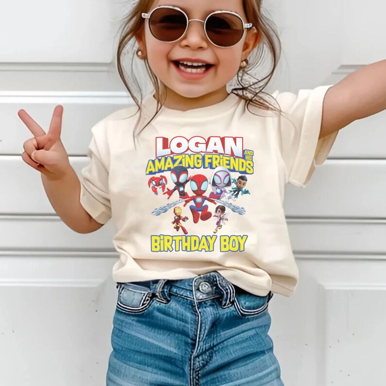 May include: A cream-colored t-shirt featuring the text "LOGAN AND HIS AMAZING FRIENDS" above a graphic of several cartoon superheroes. Below the graphic is the text "BIRTHDAY BOY" in yellow. The shirt is worn by a child.