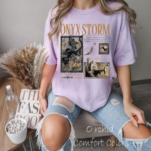 May include: A light purple t-shirt with the text "ONYX STORM" in gold lettering. The shirt features a design of postage stamps with fantasy illustrations, including a dragon and a castle. The words "Orchid Comfort Colors" are printed at the bottom.