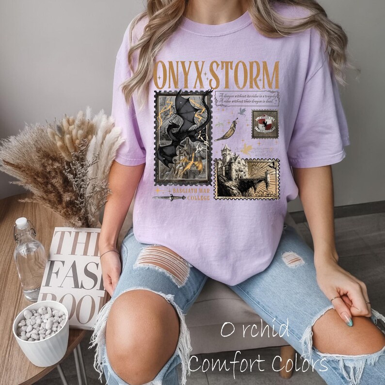 May include: A light purple t-shirt with the text "ONYX STORM" in gold lettering. The shirt features a design of postage stamps with fantasy illustrations, including a dragon and a castle. The words "Orchid Comfort Colors" are printed at the bottom.