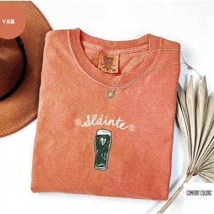 K&ouml;nnte beinhalten: Ein korallenfarbenes T-Shirt mit dem Wort "Sl&aacute;inte" und einer Abbildung eines Pints Stout. Das Shirt hat einen Rundhalsausschnitt und ein Etikett mit der Aufschrift "Comfort Colors". Eine goldene Halskette und ein brauner Hut sind ebenfalls im Bild.