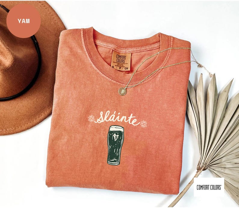 K&ouml;nnte beinhalten: Ein korallenfarbenes T-Shirt mit dem Wort "Sl&aacute;inte" und einer Abbildung eines Pints Stout. Das Shirt hat einen Rundhalsausschnitt und ein Etikett mit der Aufschrift "Comfort Colors". Eine goldene Halskette und ein brauner Hut sind ebenfalls im Bild.
