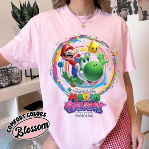 May include: A light pink t-shirt featuring a colorful graphic of Mario, Yoshi, and a star from the Super Mario Galaxy movie. The shirt also has the text "The Super Mario Galaxy Movie" and "Comfort Colors Blossom".
