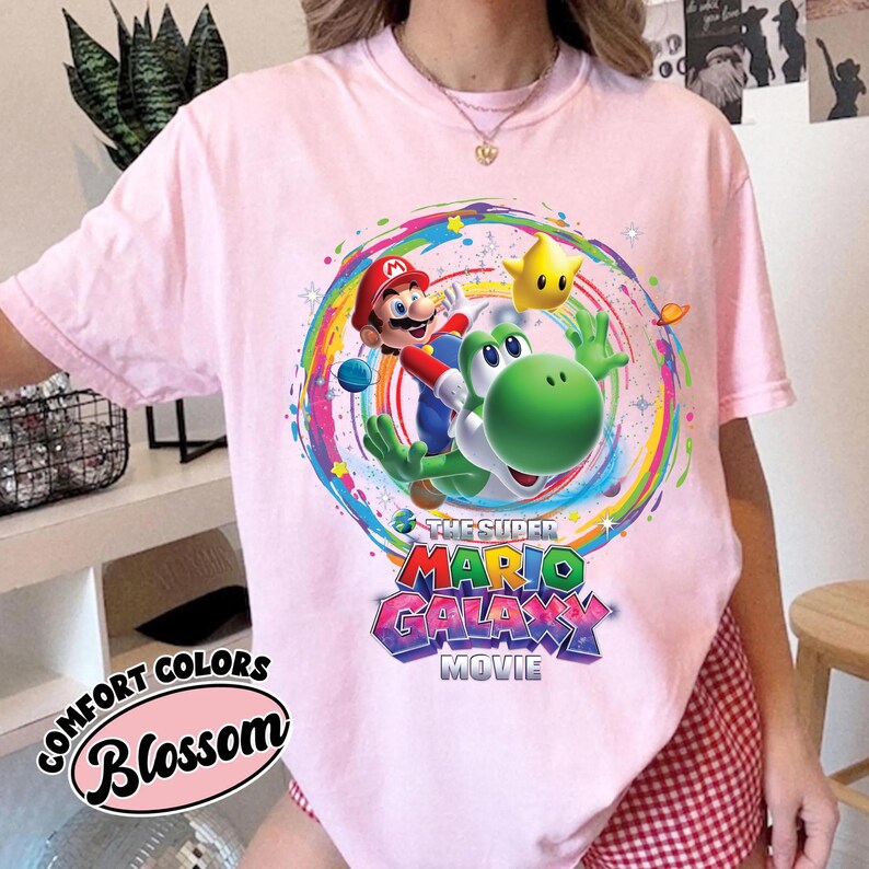 May include: A light pink t-shirt featuring a colorful graphic of Mario, Yoshi, and a star from the Super Mario Galaxy movie. The shirt also has the text "The Super Mario Galaxy Movie" and "Comfort Colors Blossom".