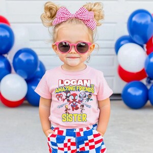 May include: A young child wearing a pink t-shirt with the text "LOGAN AND AMAZING FRIENDS SISTER" and a graphic of superheroes. The child is also wearing pink sunglasses and a pink and white checkered bow. Blue, white, and red balloons are in the background.