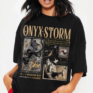 May include: Black t-shirt featuring a fantasy-themed design. The design includes the text "ONYX STORM" in gold lettering, along with illustrations of dragons, castles, and the text "BASGIATH WAR COLLEGE". The shirt has a crew neck and short sleeves.