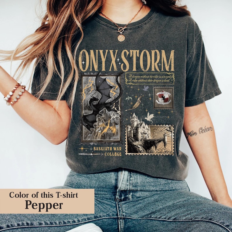 May include: Pepper-colored t-shirt with the text "ONYX STORM" in gold lettering. The shirt features a graphic of a black dragon, a castle, and the words "BASGIATH WAR COLLEGE". The shirt also has the quote "A dragon without its rider is a tragedy. A rider without their dragon is dead."