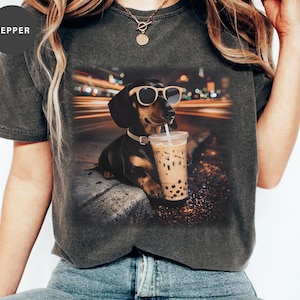 May include: Dark gray t-shirt featuring a dachshund wearing sunglasses and drinking a beverage with a straw. The dog is sitting on a curb with a blurred city background. The word "PEPPER" is on the upper left of the shirt.