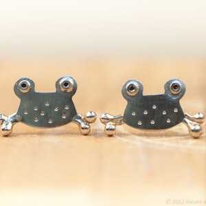 May include: A pair of silver frog earrings with red gemstone eyes. The frogs are detailed with small dots and have a slightly rounded shape.
