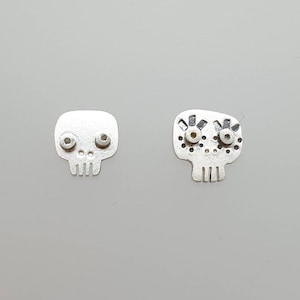 May include: A pair of silver skull earrings. One skull has simple eyes, while the other has more detailed eyes with eyelashes.