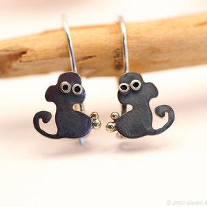 May include: A pair of silver monkey earrings with black eyes and gold accents. The monkeys are facing forward with their tails curled behind them.