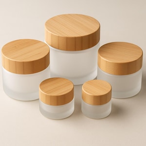 Puede incluir: Cinco tarros de vidrio esmerilado con tapas de bambú de diferentes tamaños. Los tarros son cilíndricos y tienen un acabado liso y mate. Las tapas son redondas y de bambú de color claro. Los tarros están sobre un fondo neutro.