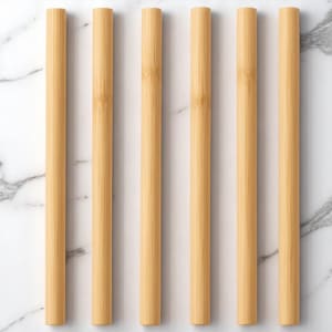 Può includere: Sei cannucce cilindriche di bambù disposte verticalmente su uno sfondo di marmo bianco. Le cannucce sono di colore marrone chiaro, con la venatura del bambù visibile. L'immagine è ben illuminata, evidenziando la consistenza e il colore naturali delle cannucce.