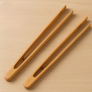 Puede incluir: Dos pinzas de cocina de bambú marrón claro. Las pinzas tienen un diseño largo y delgado con una punta redondeada y un recorte en el mango. Están sobre una superficie de madera marrón claro. Las pinzas miden unos 30 cm de largo.