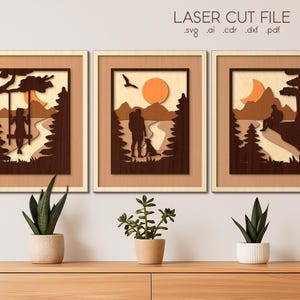 Könnte beinhalten: Drei gerahmte Laser-Cut-Holzkunstwerke, die Szenen im Freien darstellen. Jedes zeigt eine Silhouette einer Person oder von Personen in einer natürlichen Umgebung mit Bäumen, einer Sonne oder einem Mond und einem Pfad. Der Text "LASER CUT FILE" steht oben.
