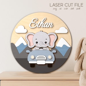 Può includere: Decorazione murale circolare in legno con un elefante dei cartoni animati alla guida di un'auto, con il nome "Ethan" sopra. Il design include montagne, nuvole e il testo "LASER CUT FILE". La tavolozza dei colori include azzurro, grigio e beige.