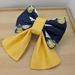 May include: A handmade bow tie with a navy blue bee print fabric. The bow's centre and tails are a solid yellow. The bow tie is likely for a pet or child.