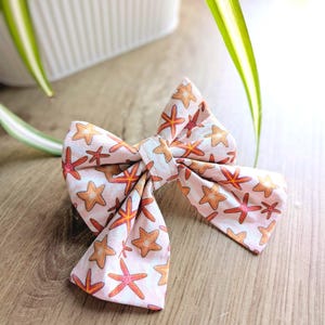 May include: A fabric bow tie with a pattern of orange and red starfish on a pale pink fabric. The bow tie is knotted in the middle and rests on a wooden surface.